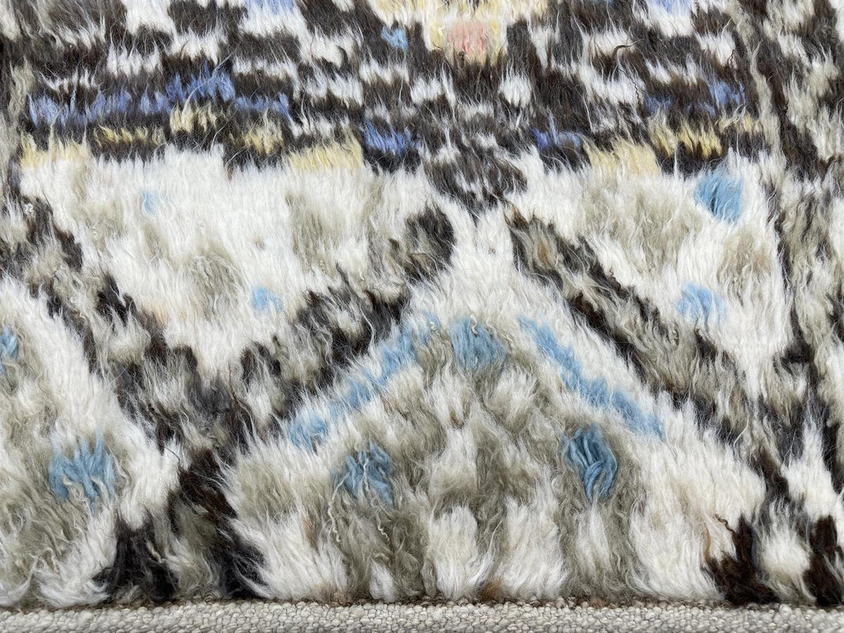 Close-up of a textured, shaggy rug with an abstract pattern in cream, gray, black, beige, and light blue. Atlantica Organics curates antique and handmade rugs, Persian, Afghan, Tibetan, Pakistani, Turkish Oushak, Kilim, Khotan, tribal and contemporary textiles for luxury interiors, homes and real estate. Our collections connect timeless craftsmanship with interior design, furniture, fine art, architecture and modern living. Explore curated rugs, collectible textiles, natural materials, cultural heritage, su