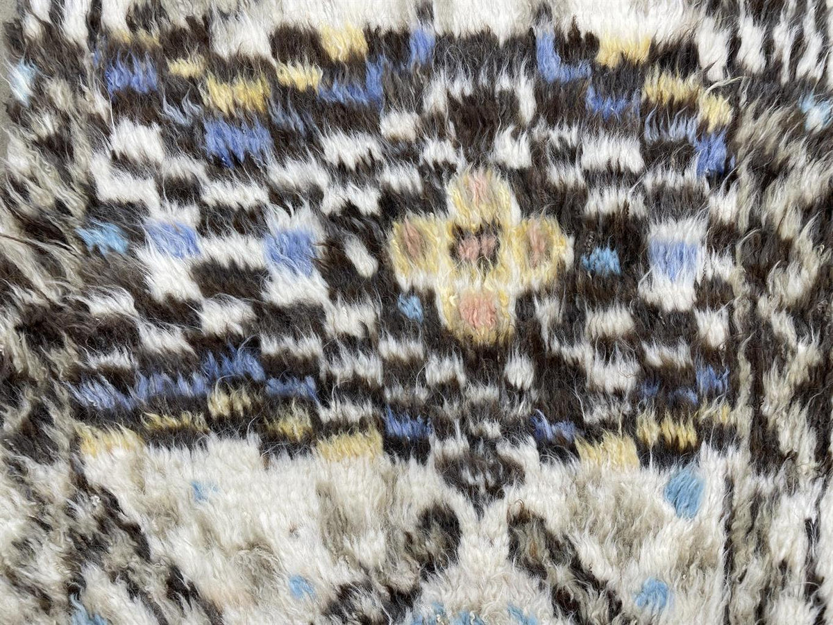 Close-up of a textured, shaggy area rug with a vintage-style geometric and floral pattern in cream, brown, black, blue, and yellow. Atlantica Organics curates antique and handmade rugs, Persian, Afghan, Tibetan, Pakistani, Turkish Oushak, Kilim, Khotan, tribal and contemporary textiles for luxury interiors, homes and real estate. Our collections connect timeless craftsmanship with interior design, furniture, fine art, architecture and modern living. Explore curated rugs, collectible textiles, natural materi