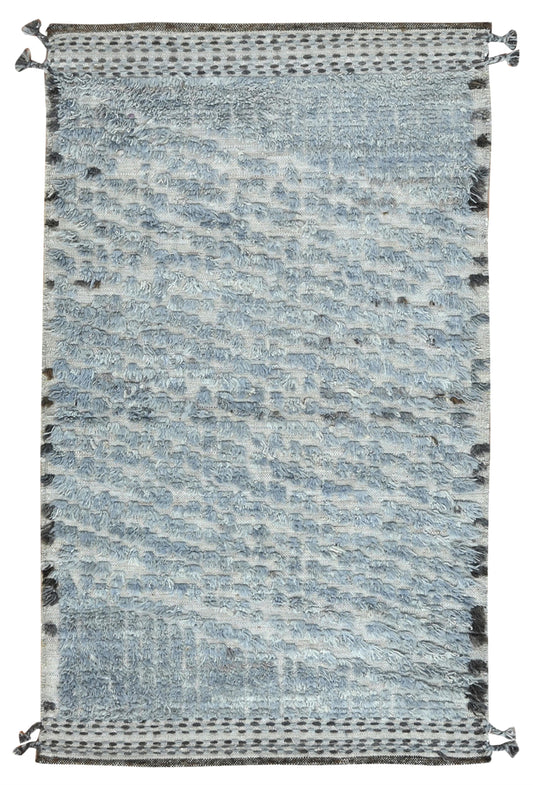A rectangular woven area rug with a textured, abstract pattern in shades of light blue, gray, and white, featuring dark gray decorative borders and tassels on the top and bottom edges. Atlantica Organics curates antique and handmade rugs, Persian, Afghan, Tibetan, Pakistani, Turkish Oushak, Kilim, Khotan, tribal and contemporary textiles for luxury interiors, homes and real estate. Our collections connect timeless craftsmanship with interior design, furniture, fine art, architecture and modern living. Explo