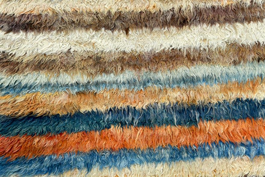 A close-up of a shaggy, textured rug with horizontal stripes in cream, brown, light blue, orange, and dark blue. Atlantica Organics curates antique and handmade rugs, Persian, Afghan, Tibetan, Pakistani, Turkish Oushak, Kilim, Khotan, tribal and contemporary textiles for luxury interiors, homes and real estate. Our collections connect timeless craftsmanship with interior design, furniture, fine art, architecture and modern living. Explore curated rugs, collectible textiles, natural materials, cultural herit
