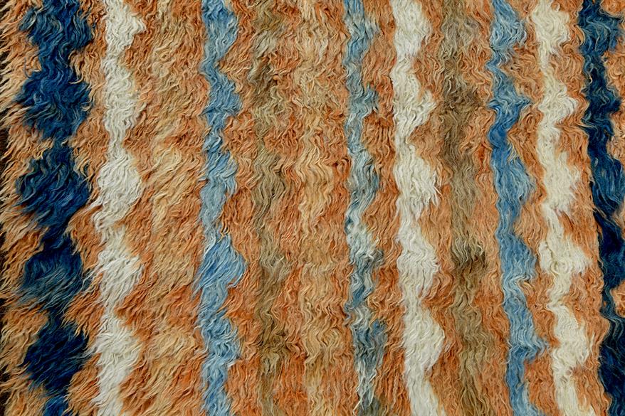 A close-up of a shaggy, textured rug or fabric with vertical zigzag stripes in blue, cream, tan, and brown. Atlantica Organics curates antique and handmade rugs, Persian, Afghan, Tibetan, Pakistani, Turkish Oushak, Kilim, Khotan, tribal and contemporary textiles for luxury interiors, homes and real estate. Our collections connect timeless craftsmanship with interior design, furniture, fine art, architecture and modern living. Explore curated rugs, collectible textiles, natural materials, cultural heritage, 